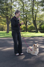 Vrouw staat op een plein tussen de bomen haar lipstick bij te werken terwijl haar hond aan de multifunctionele handsfreelijn van het merk punq op de grond naast haar ligt.