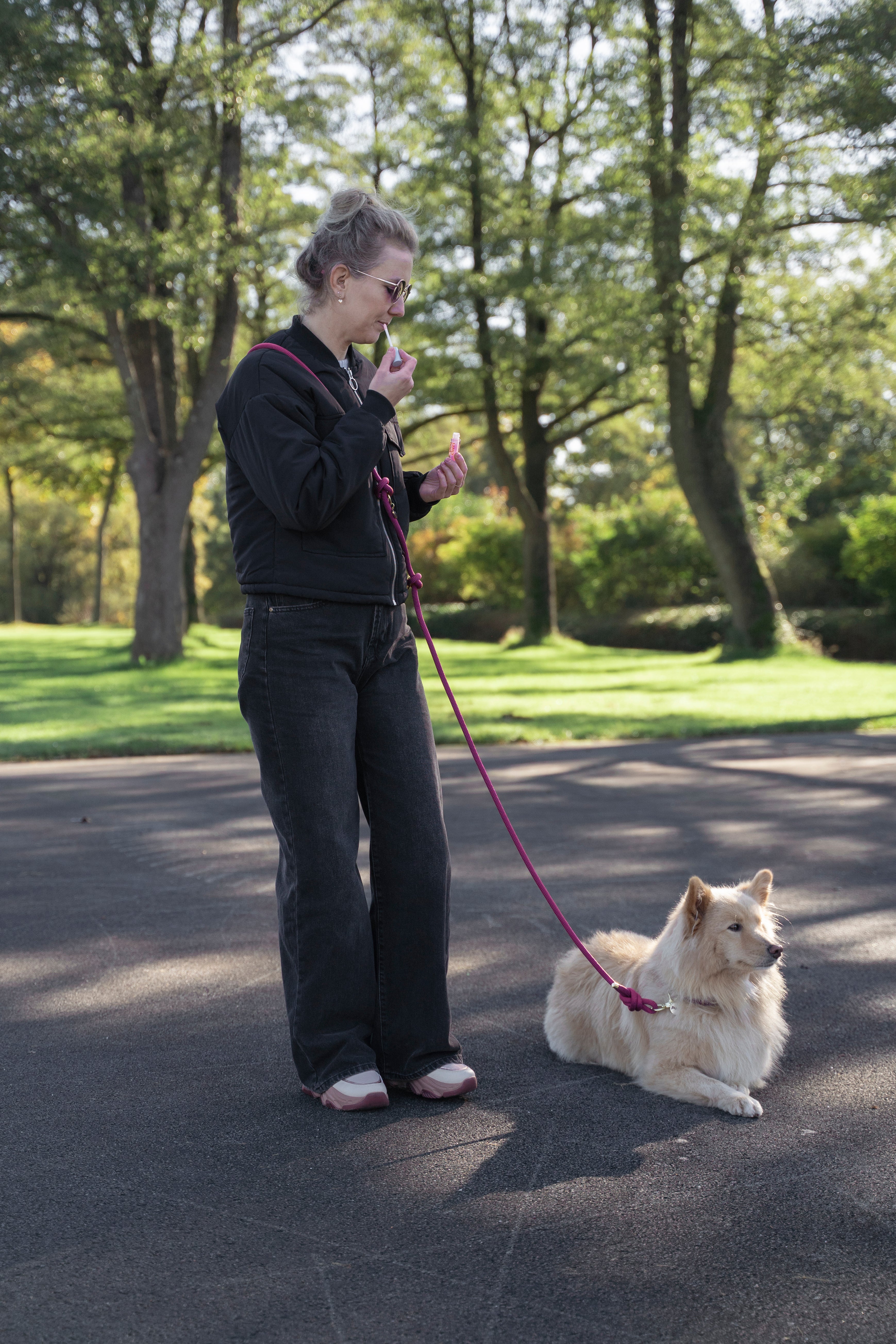 Vrouw staat op een plein tussen de bomen haar lipstick bij te werken terwijl haar hond aan de multifunctionele handsfreelijn van het merk punq op de grond naast haar ligt.