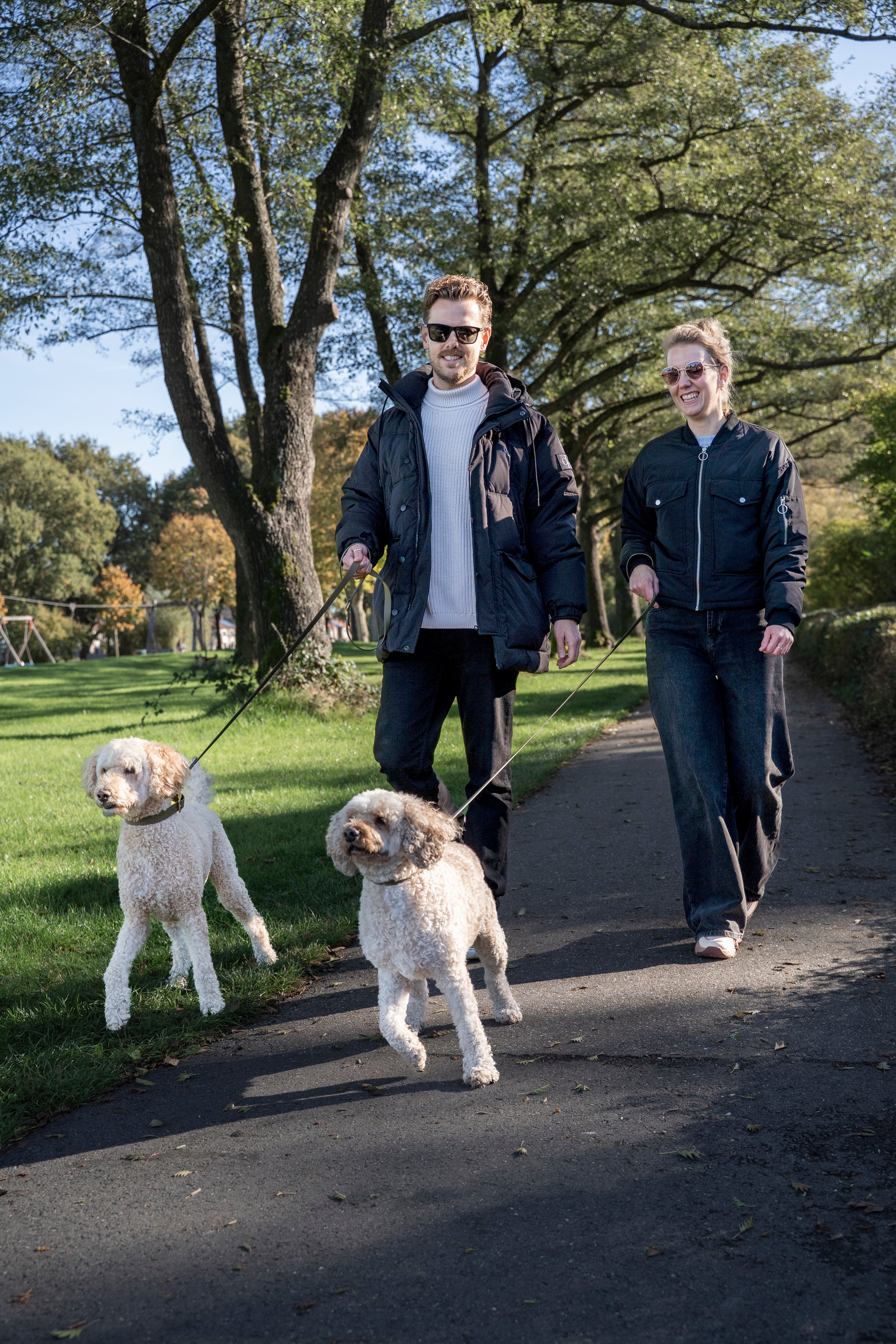 een man en vrouw lopen naast elkaar met 2 labradoodles. De honden dragen de olive green halsbanden en riemen van punq