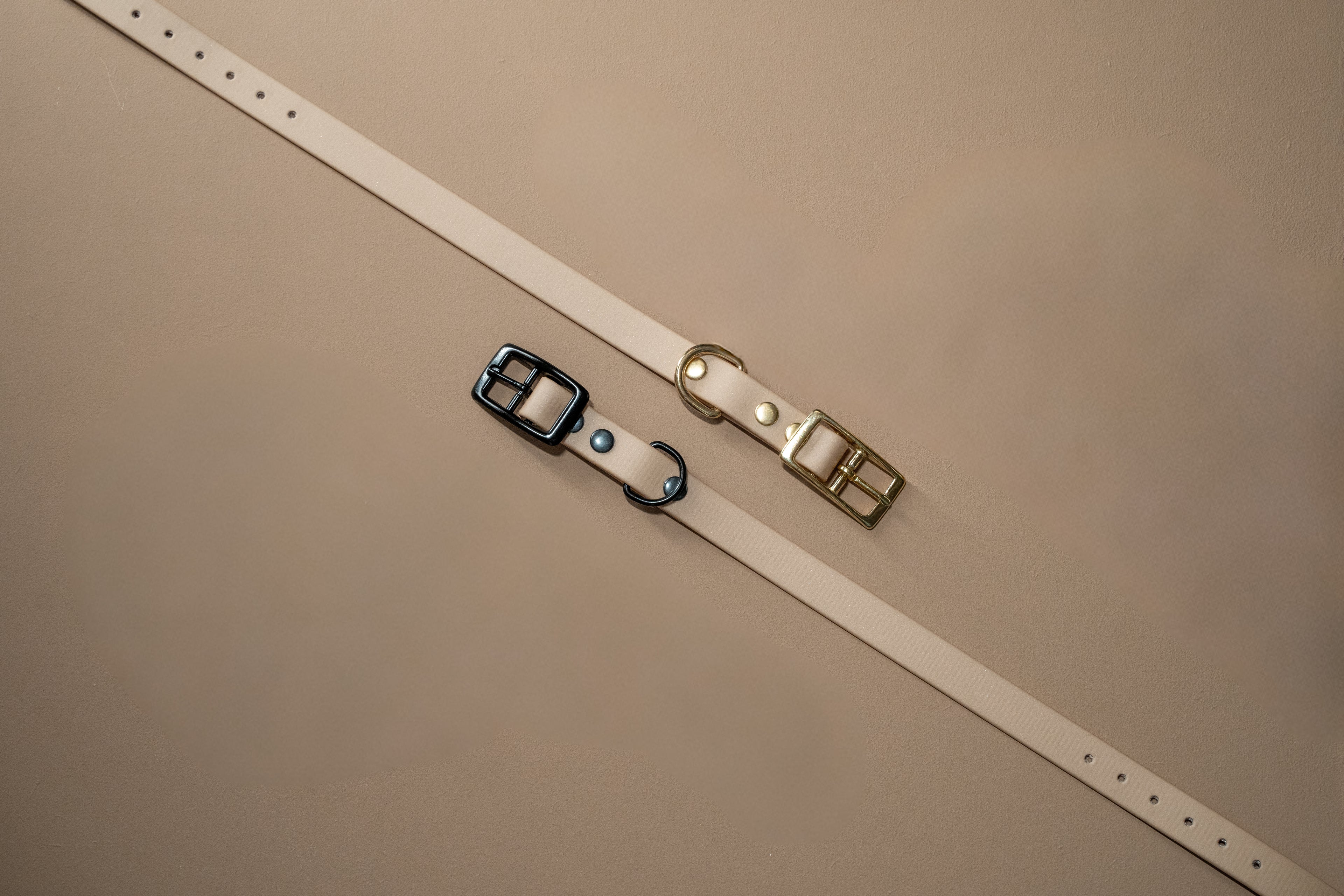 2 beige halsbanden voor een hond van bovenaf op een beige achtergrond. 1 halsband met zwarte sluiting en 1 met messing sluiting